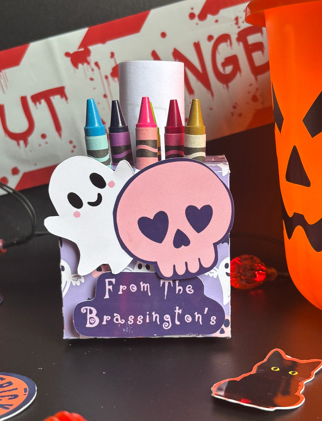 A Halloween-themed crayon holder box with a ghost and skull design, holding six crayons, set on a dark tabletop next to an orange jack-o'-lantern bucket and holiday decorations.