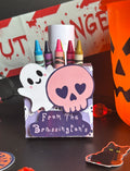 A Halloween-themed crayon holder box with a ghost and skull design, holding six crayons, set on a dark tabletop next to an orange jack-o'-lantern bucket and holiday decorations.